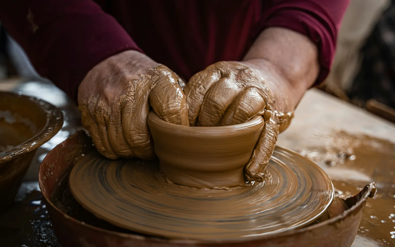 Clay-covered hands shaping a small bowl on a pottery wheel
